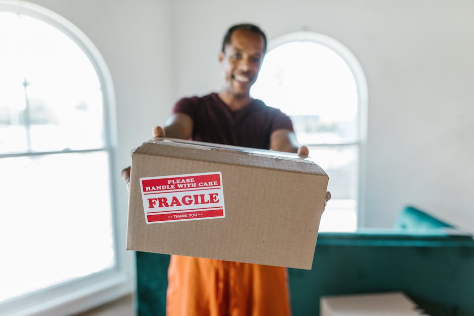 A person wearing a maroon shirt is holding an open cardboard box labeled with a red and white 'Fragile' sticker, indicating careful handling. The individual’s hands are visible, with nails painted i