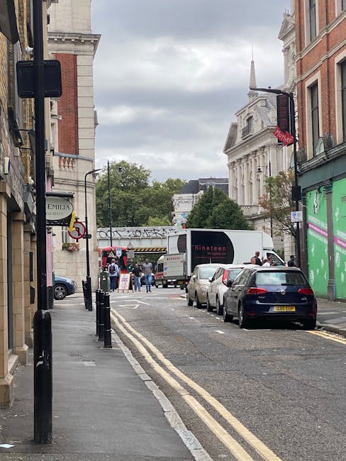 A street scene in Stockwell, London, showing a narrow road with parked cars along the right side and a loading area at the curb where Stockwell Movers are engaged in a home relocation process. On the left, the sidewalk features black bollards, a shop sign, and protective posts. Several large cardboard boxes and wrapped furniture are being carried or prepared for loading onto a white van visible further down the street, along with a smaller delivery truck parked behind it. Pedestrians walk nearby, some standing near the vehicles and boxes, while others continue along the pavement. The surrounding buildings include typical London architecture, with a prominent white building with columns in the background and a modern green storefront on the right. Overhead, the sky is overcast, casting diffuse lighting over the scene, which captures a typical urban furniture transport and packing process associated with house removals in the Stockwell area.